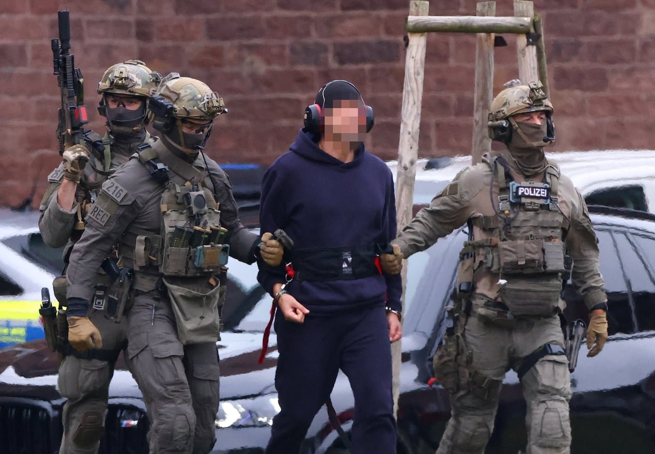 A man, obscured face, wearing headphones, a ballcap, and a hoodie, escorted by three police officers in camouflage attire with firearms.
