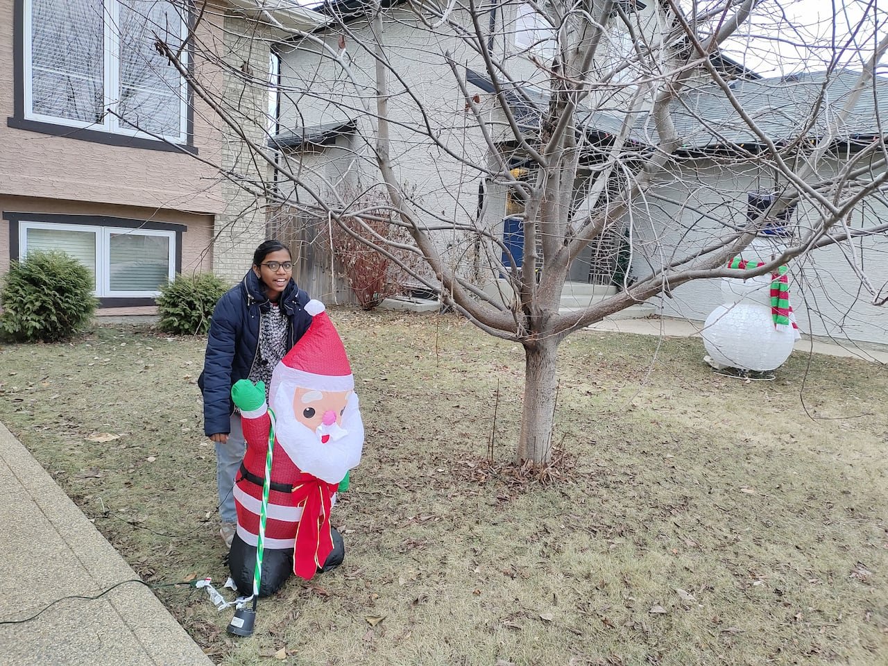 A girl in glasses stands behind an inflatable Santa Claus on a lawn devoid of snow in winter.