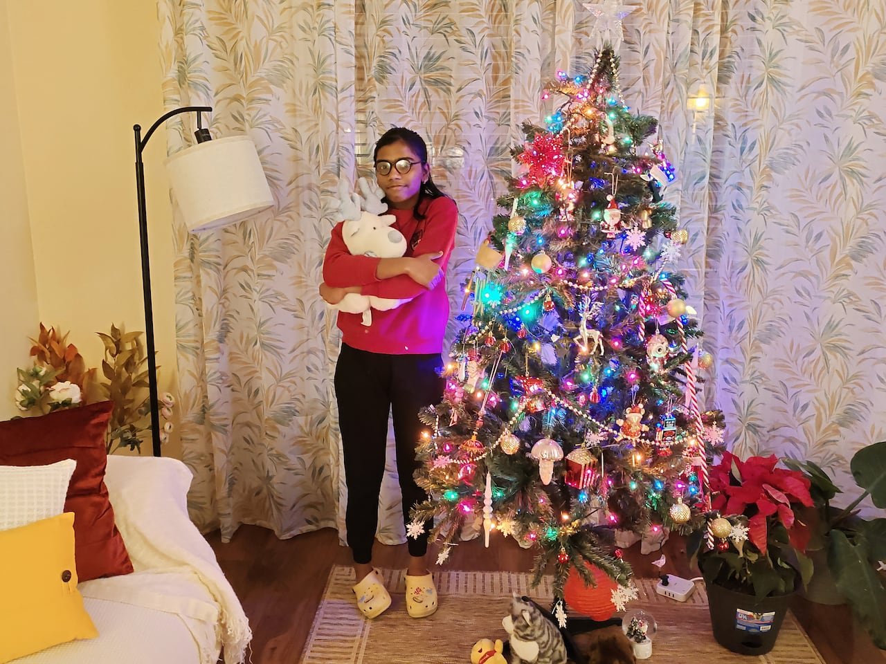A girl cuddles a stuffed animal standing beside a brightly lit Christmas tree.