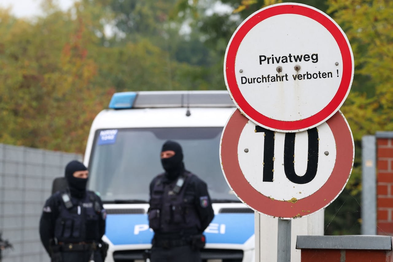 Two hooded persons in uniforms stand near a street sign.