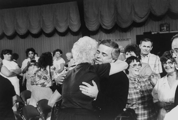 A man facing the camera hugs a woman with back to camera in a television audience.