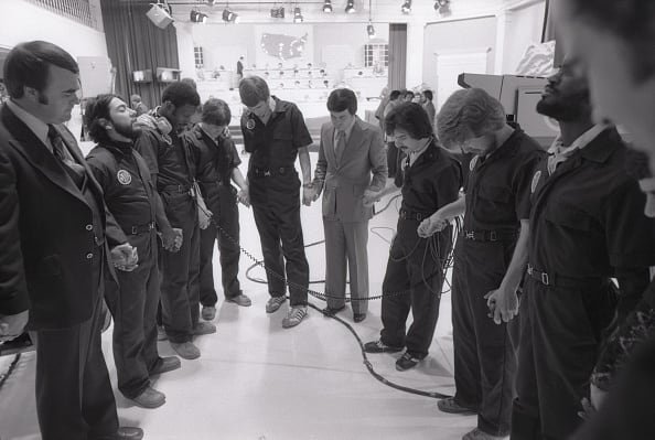 A group of people stand in a semi-circle holding hands with their heads bowed on a television set.
