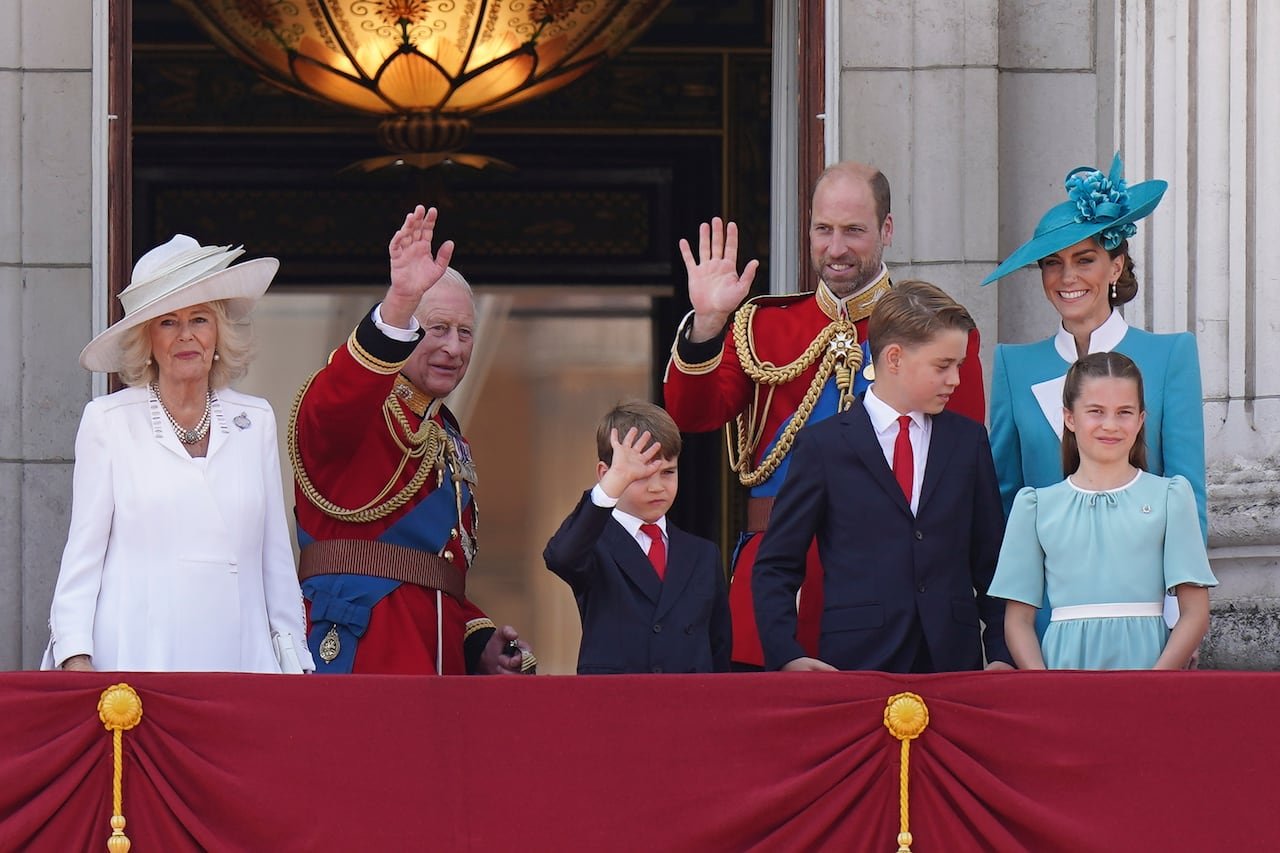 Four adults and three children wave from the balcony of a grand stone building.