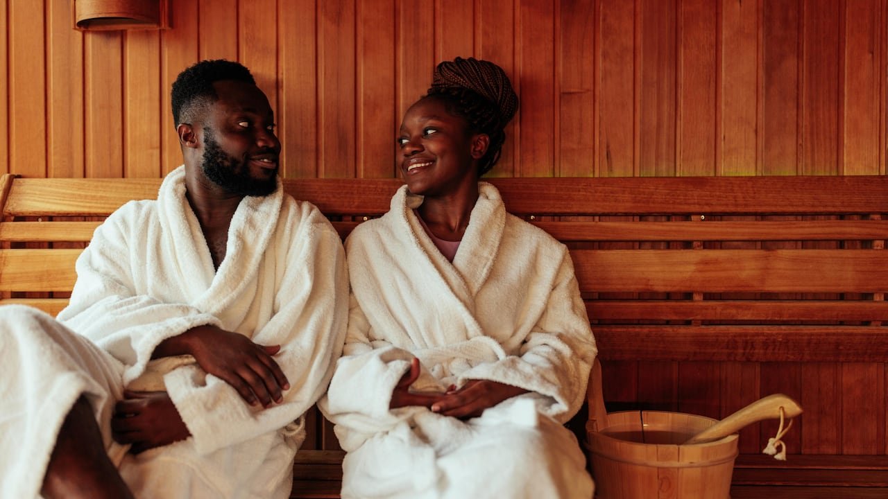 default-44 A couple in bath robes sitting on a bench in a wood-panelled sauna, smiling at each other.